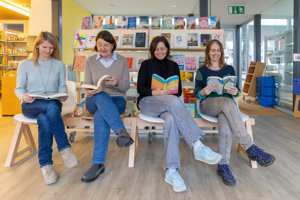 Vier Frauen sitzen in einer Reihe in einer Bibliothek und lesen ein Buch.